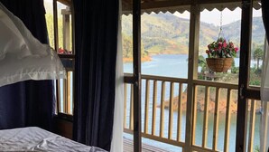 Een slaapkamer met een bed, een raam met uitzicht op een meer en heuvels, en een hangplant.