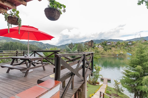 Una terraza de madera con mesas de picnic y un paraguas rojo, con vista a un lago y colinas.