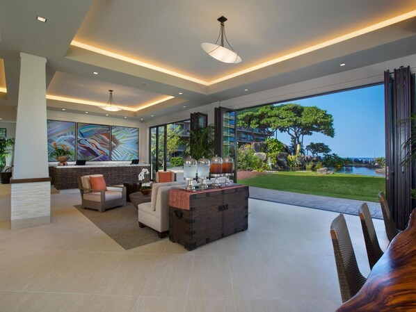A spacious living area with a large window offering a view of a landscaped garden and a body of water. There are multiple seating areas, a coffee table, and a decorative cabinet with a drink dispenser.