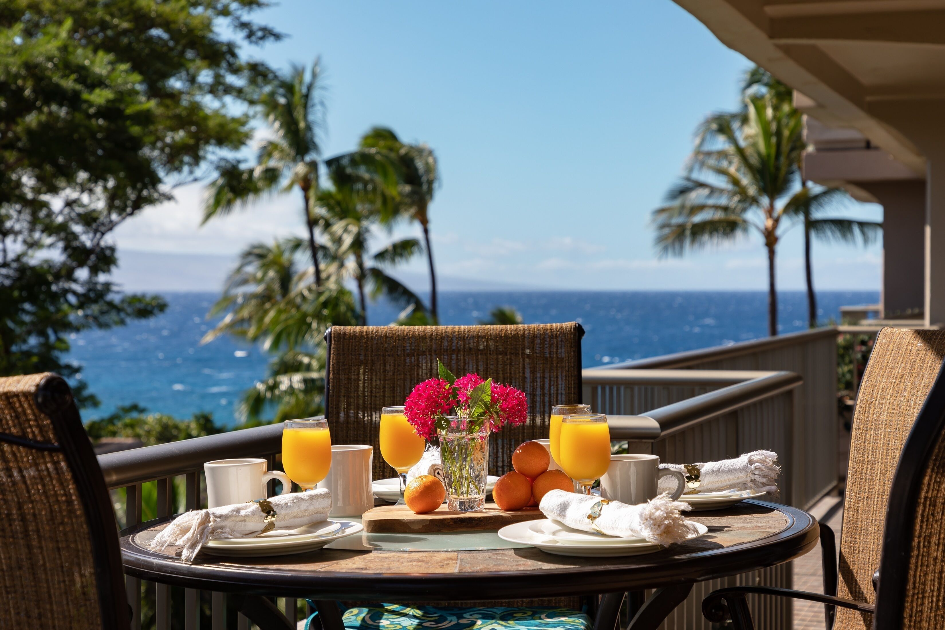 A table set with breakfast items, including orange juice, a white mug, and a plate with a napkin, overlooking a scenic ocean view with palm trees.