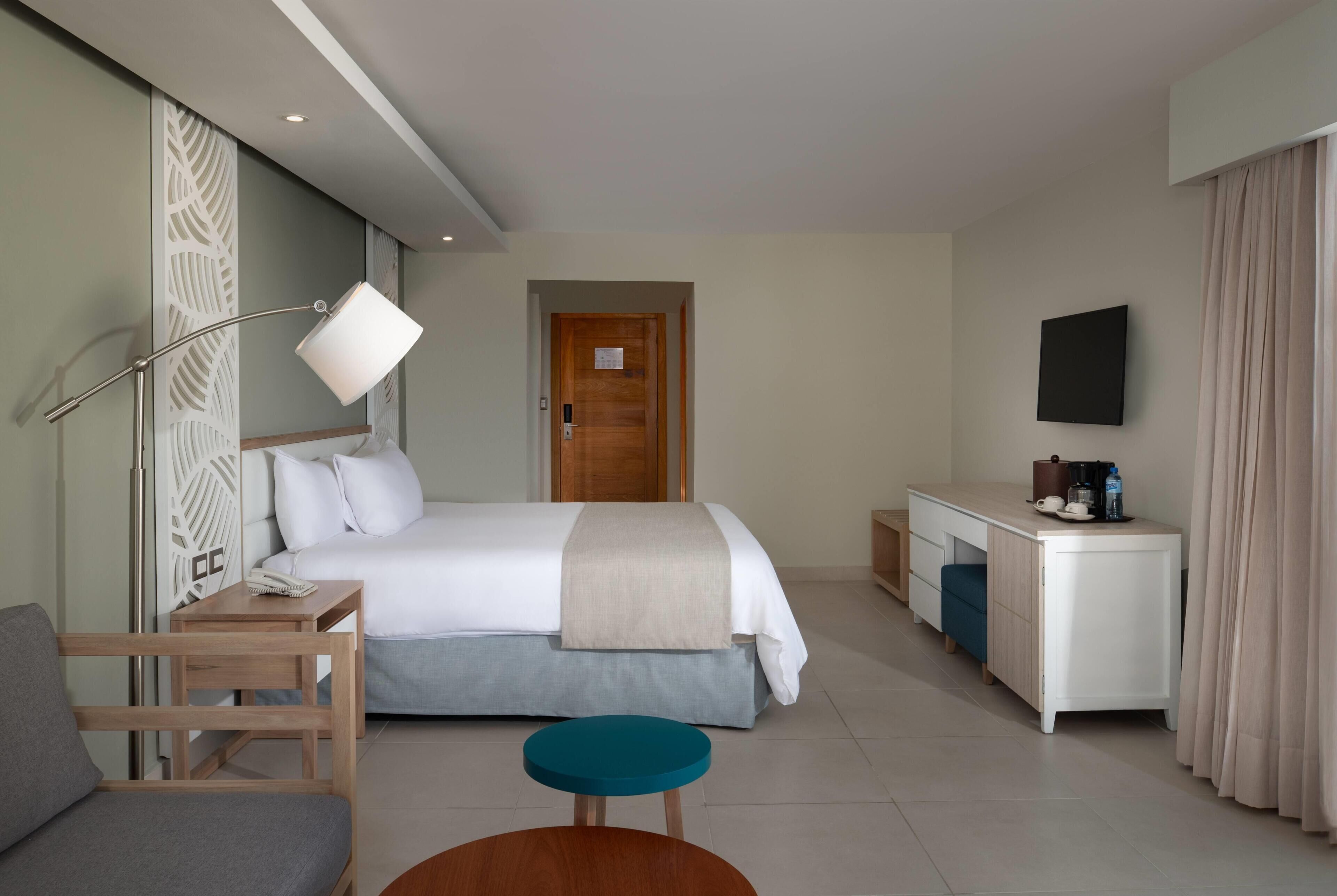 Ein Hotelzimmer mit Bett, Nachttisch, Lampe, Schreibtisch, Stuhl und einem an der Wand befestigten Fernseher.