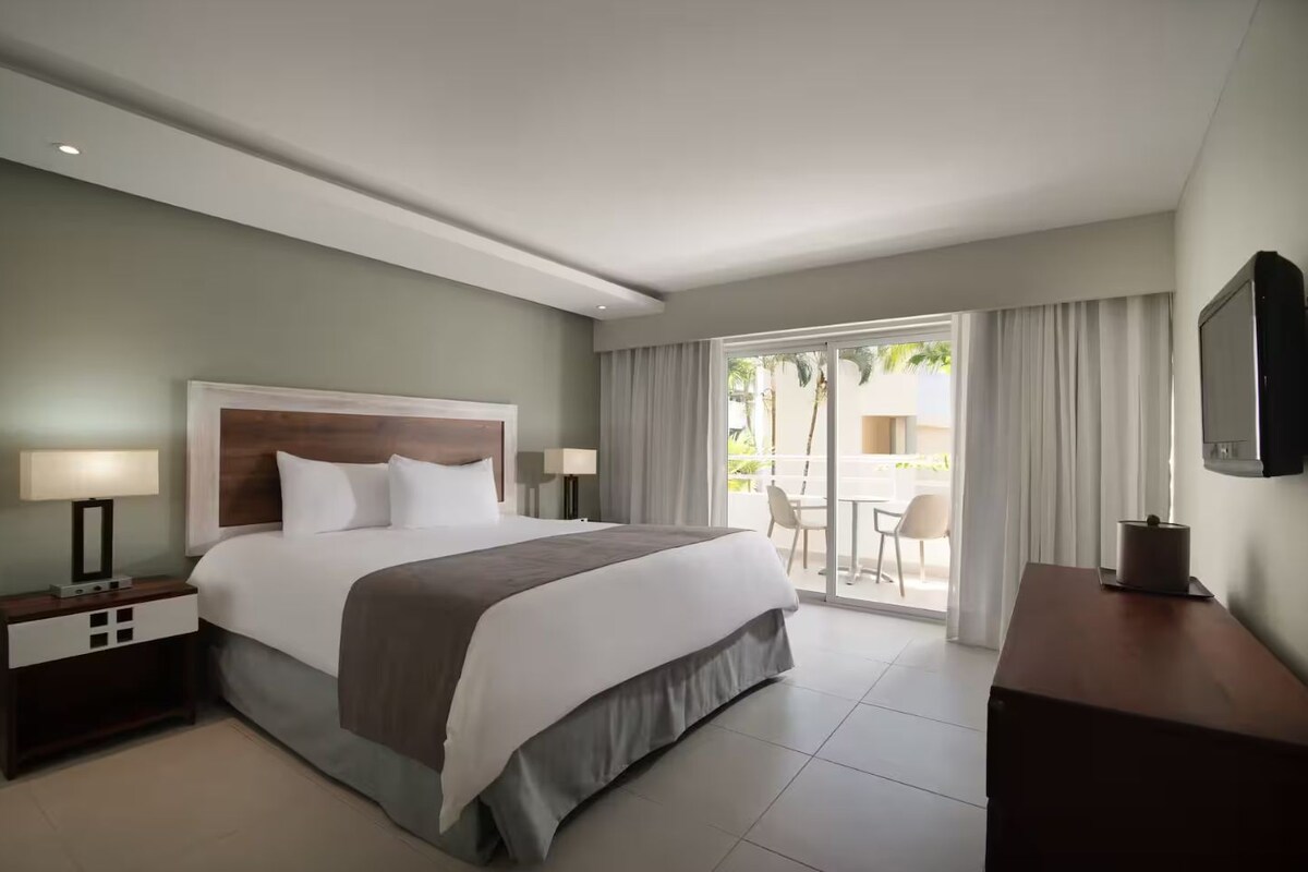 Une chambre d’hôtel avec un grand lit, deux tables de chevet avec des lampes, une télévision et un balcon équipé d’une table et de chaises.