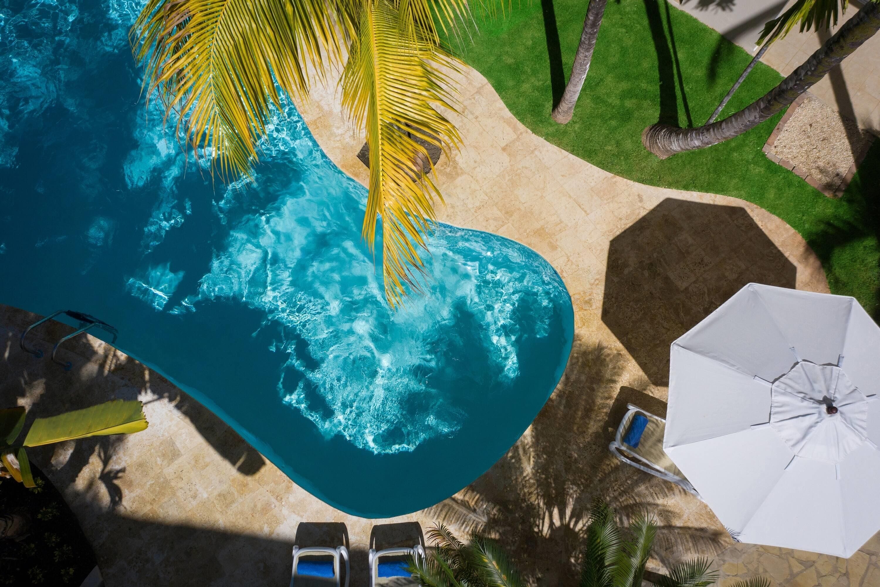 An aerial view of a swimming pool with palm trees, lounge chairs, and a white umbrella.