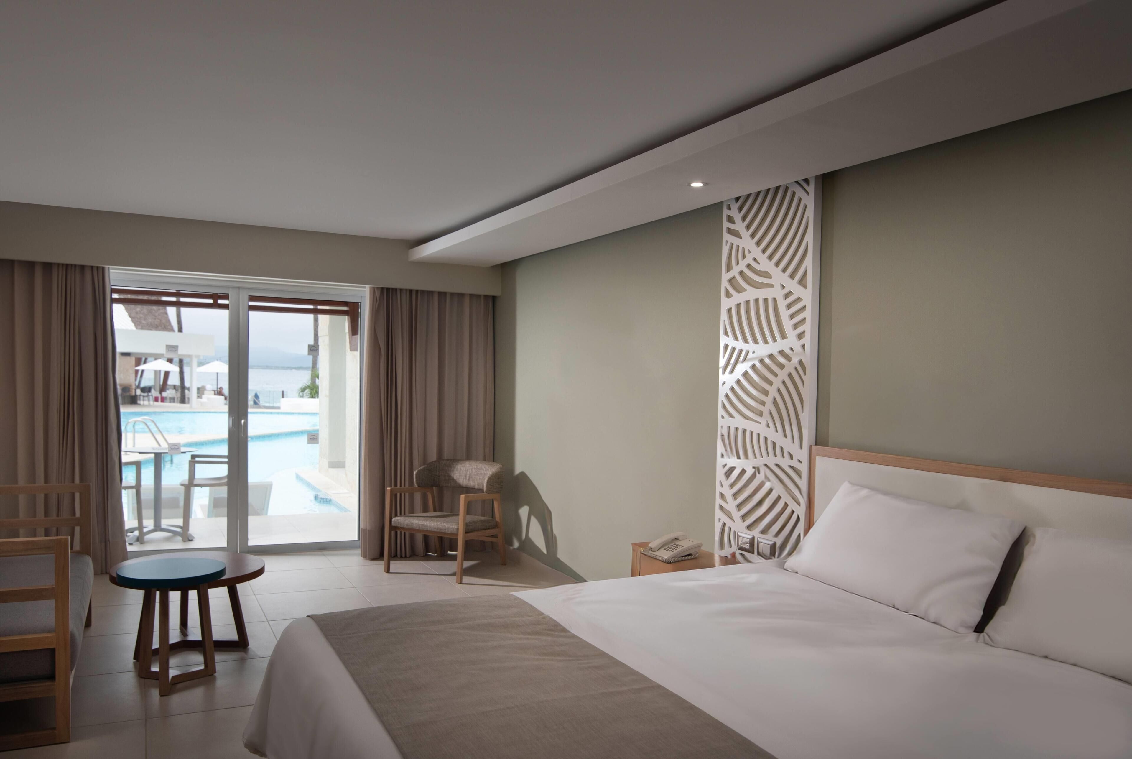 Hotelkamer met een bed, een stoel, een klein tafeltje, een telefoon en uitzicht op een zwembad door een glazen deur.