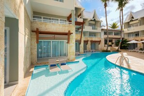 Une piscine avec des chaises longues, un espace de détente couvert et un immeuble à plusieurs étages doté de balcons.