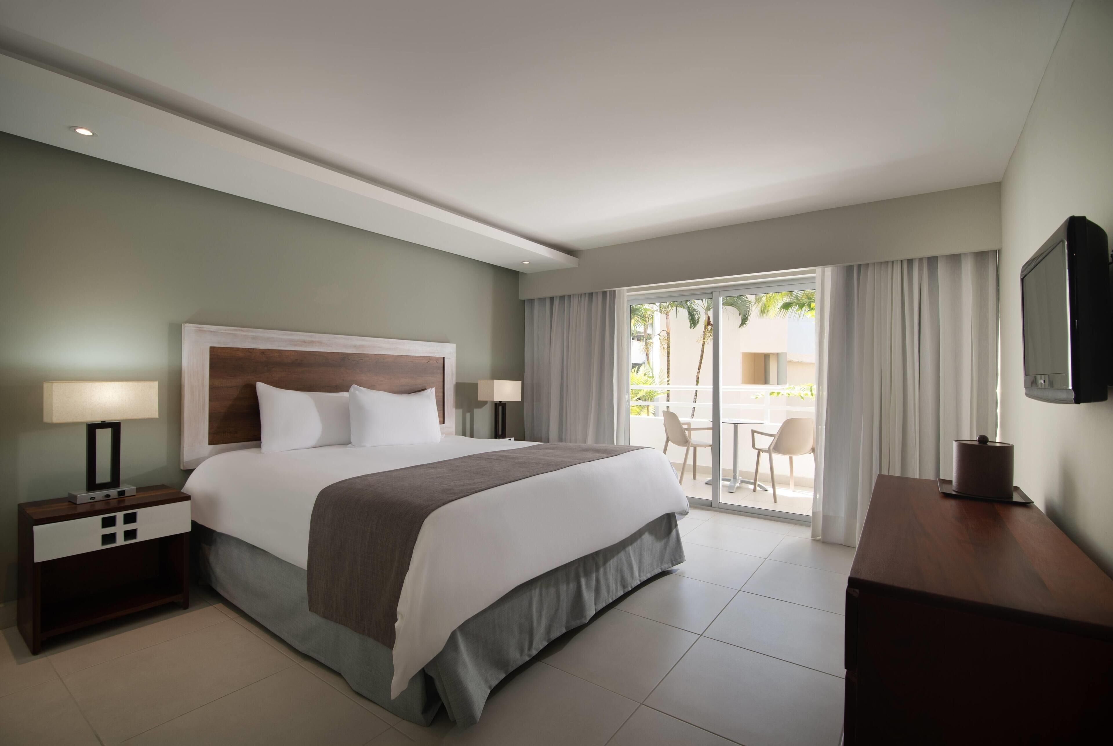 A hotel room with a bed, nightstands, a TV, a desk, and a view of the outdoors.