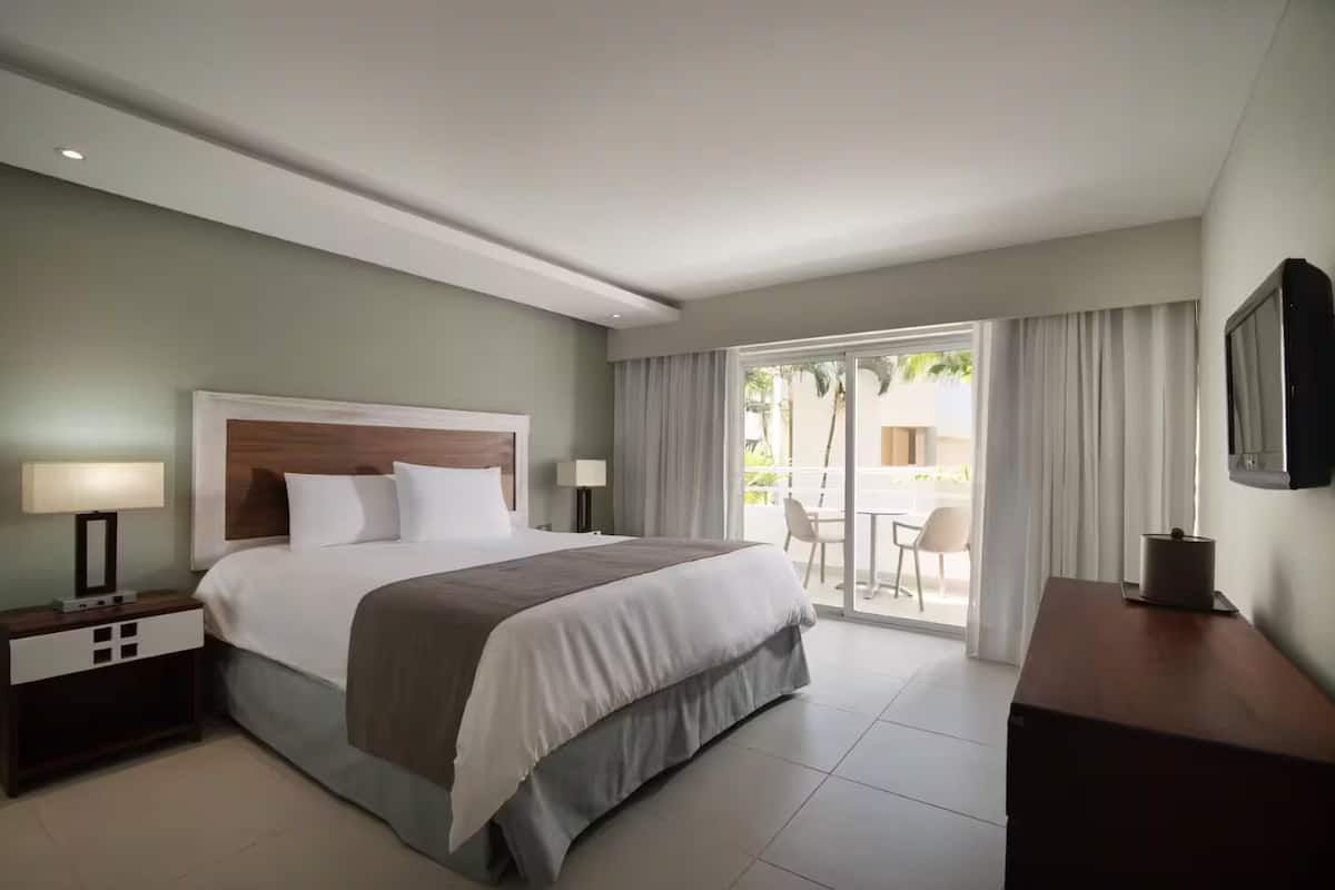 A hotel room with a bed, nightstands, a desk, a TV, and a balcony with outdoor seating.