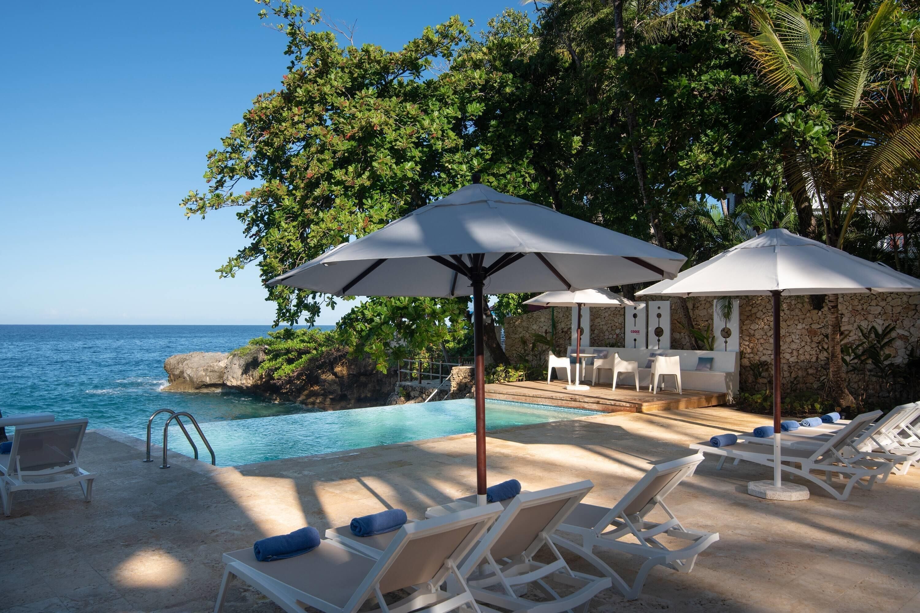 A poolside area with lounge chairs and umbrellas, a stone wall, and a view of the ocean.