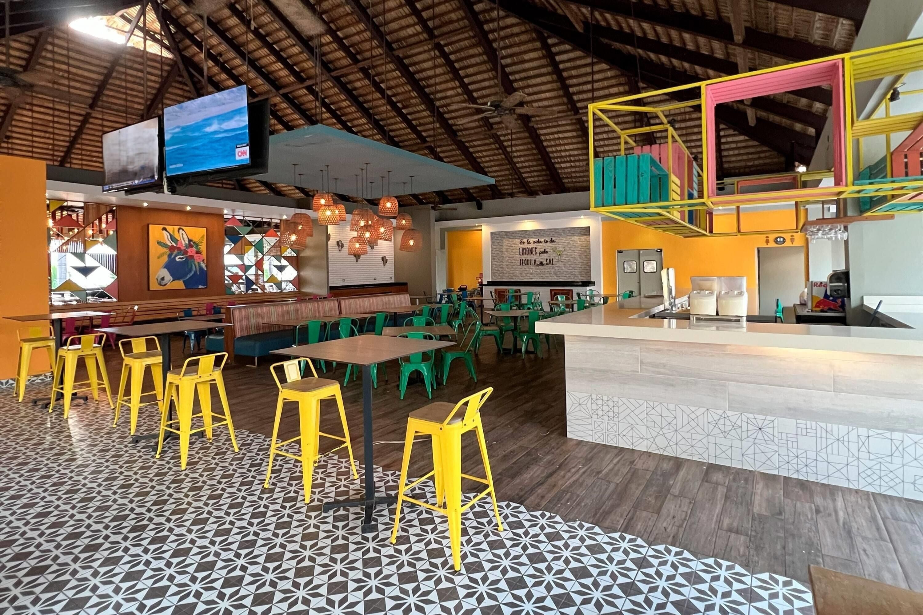 Ein Restaurant mit hohen Decken, geometrischer Wandkunst und einer Mischung aus gelben und grünen Stühlen.