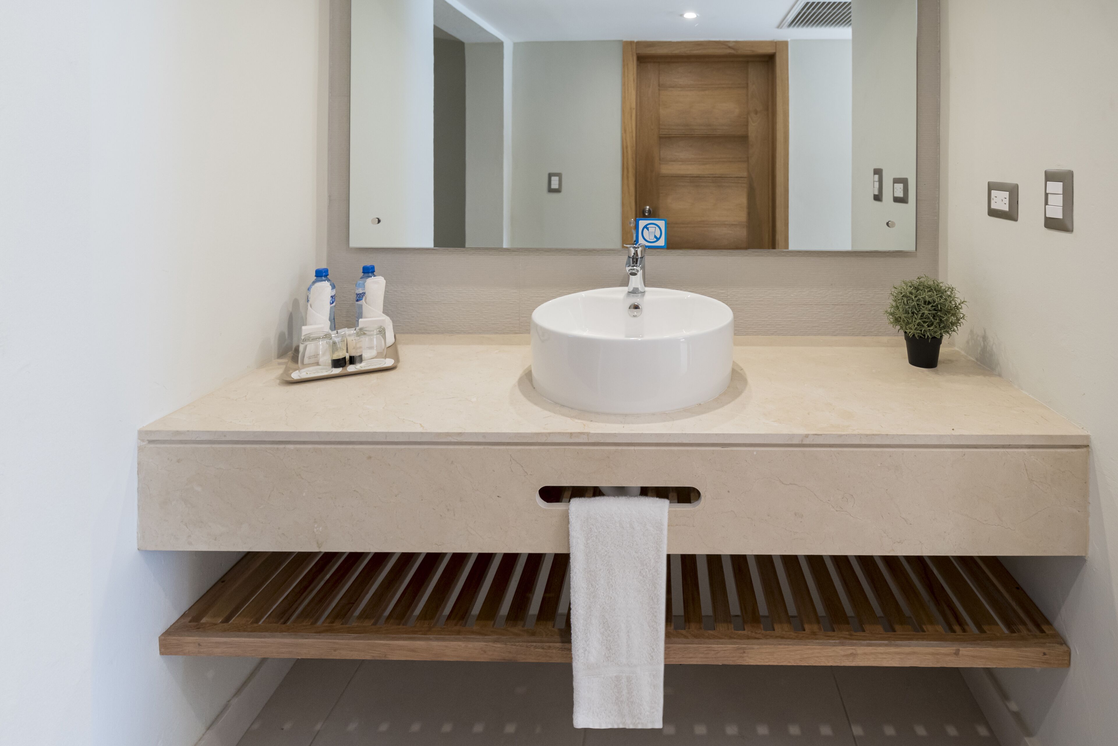 Un baño moderno con un lavamanos redondo, un espejo encima y un perchero debajo. Hay dos botellas de jabón para manos y una pequeña planta en maceta sobre la encimera.