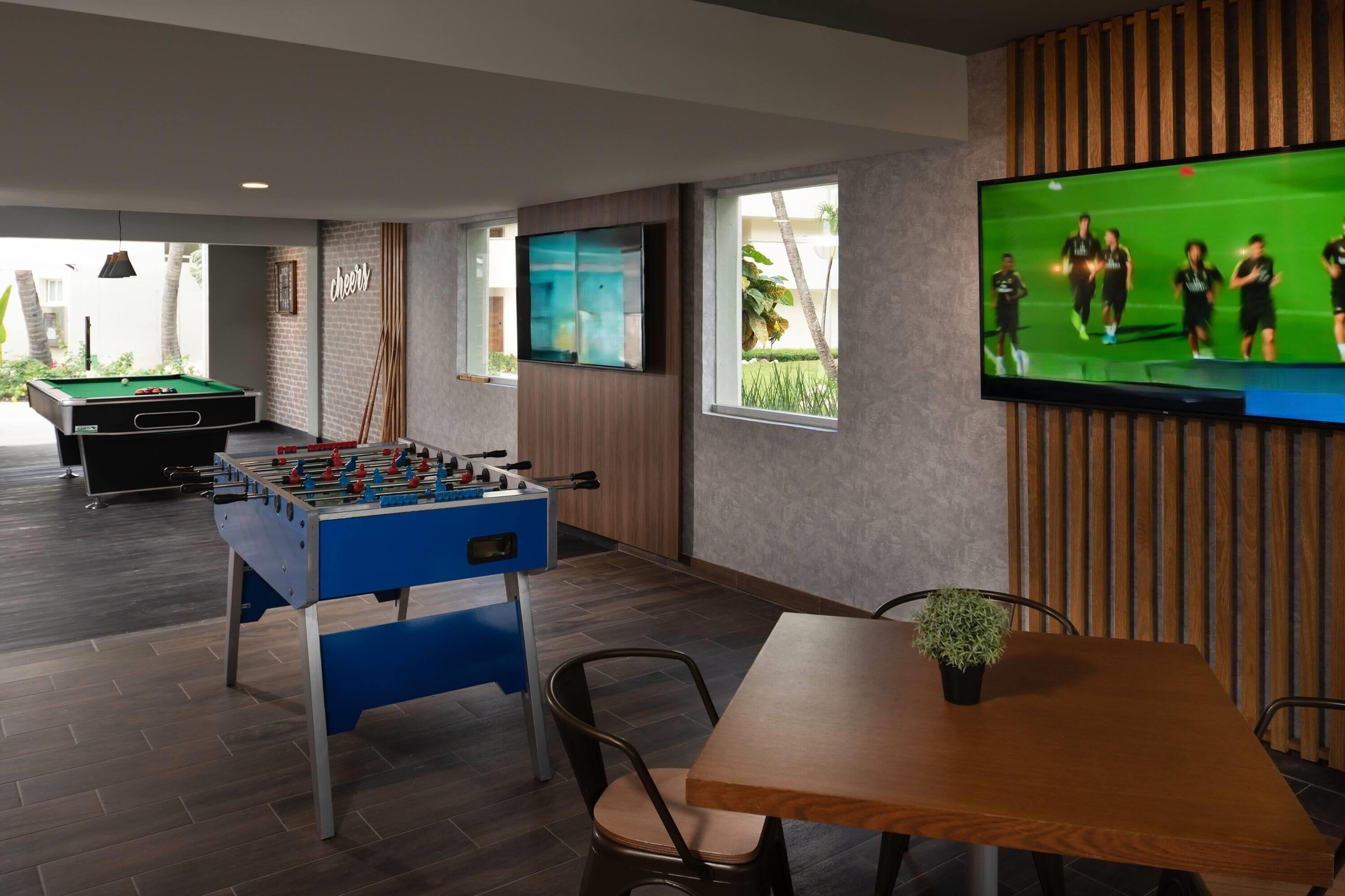 Une pièce équipée d’un baby-foot, d’une table de billard, d’un téléviseur diffusant un match de football et d’une table à manger avec des chaises.
