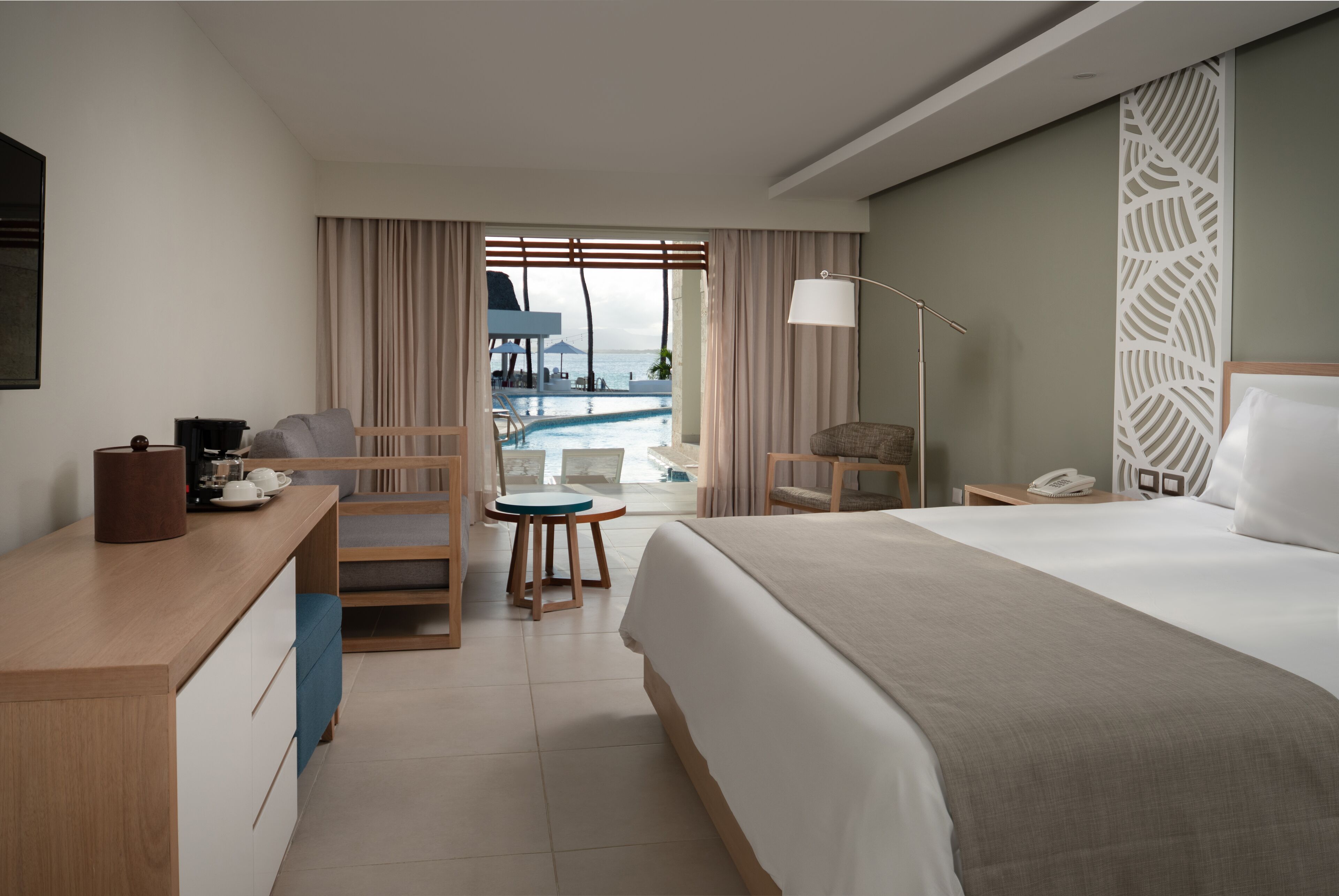 Hotelkamer met een bed, bureau met koffiezetapparaat, stoel, klein tafeltje en uitzicht op een zwembad en strand.