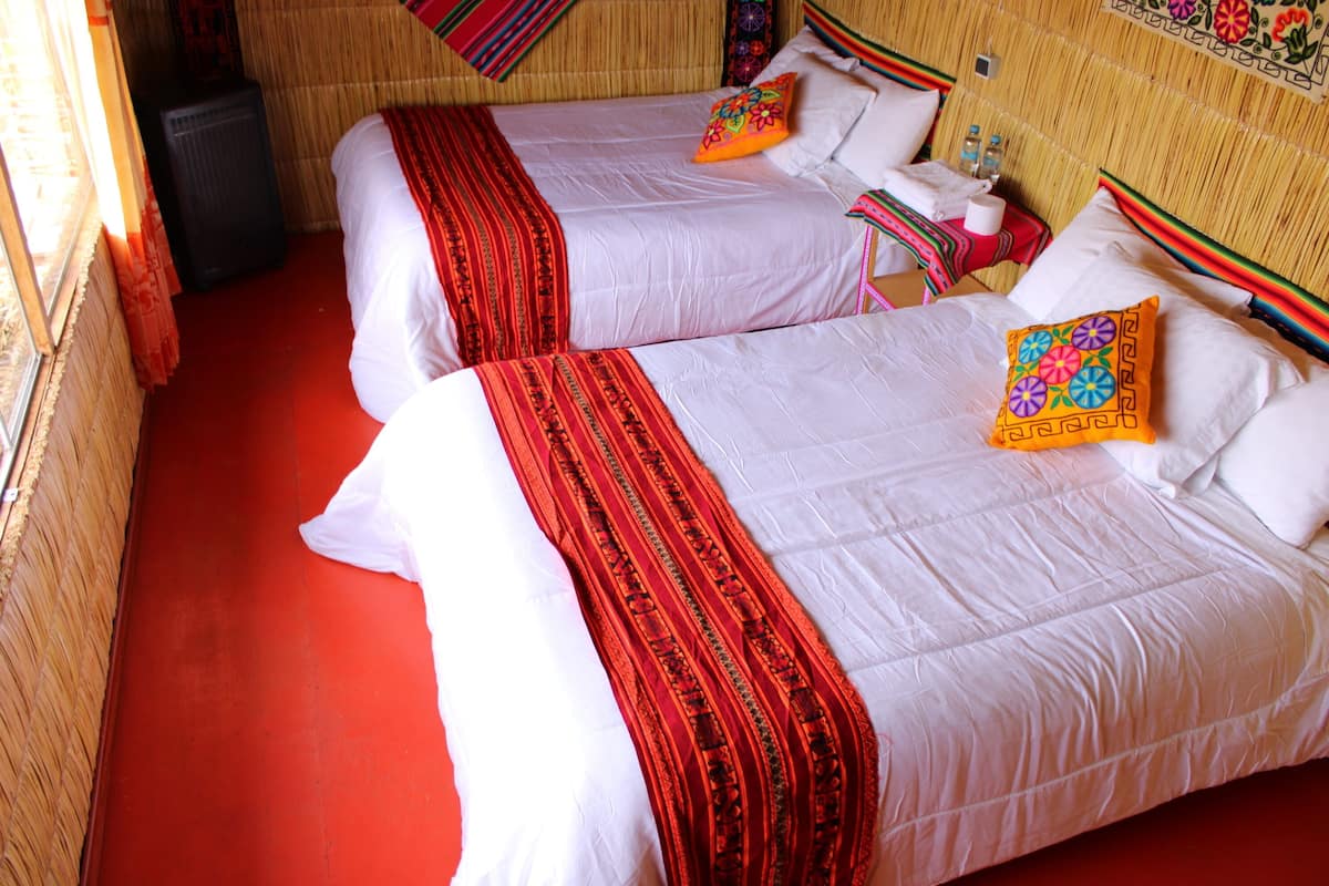 Une chambre avec deux lits, chacun agrémenté de draps blancs et de coussins décoratifs rouges. On y trouve une petite table recouverte d’une nappe rouge et une bouteille d’eau. La chambre est dotée d’un tapis rouge et d’une tête de lit en osier.