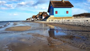 Een kustgebouw met een blauwe gevel en rode luiken, gelegen aan een kiezelstrand onder een heldere lucht.