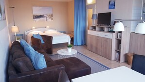Een hotelkamer met een bed, een sofa, een klein tafeltje met een plant, een televisie en een kitchenette met kastjes en een magnetron.