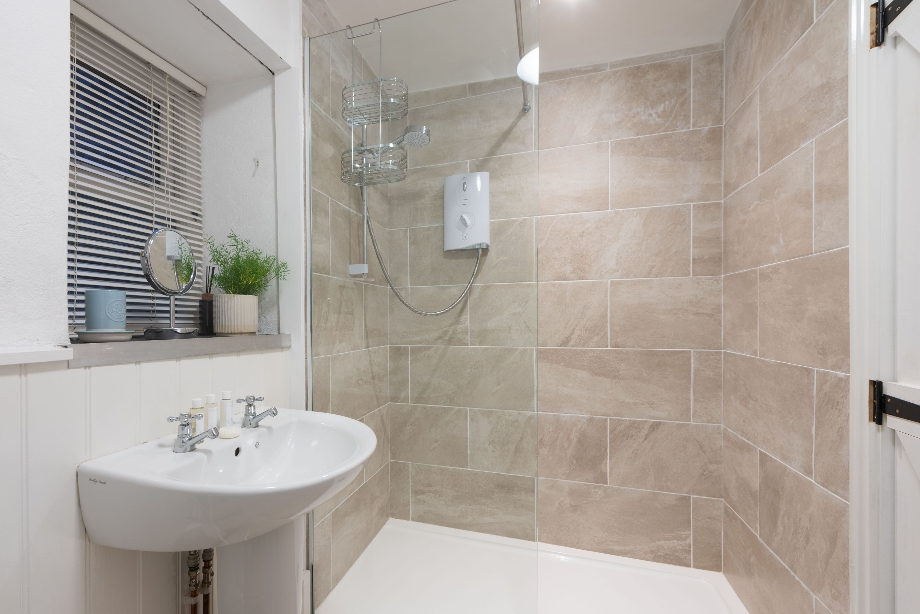 Une salle de bain avec un lavabo blanc, un miroir, une douche dotée d’une paroi en verre et des murs recouverts de carreaux beiges.
