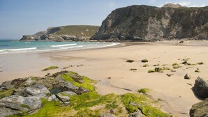 Une plage avec des affleurements rocheux, une eau d’un bleu limpide et une falaise en arrière-plan.