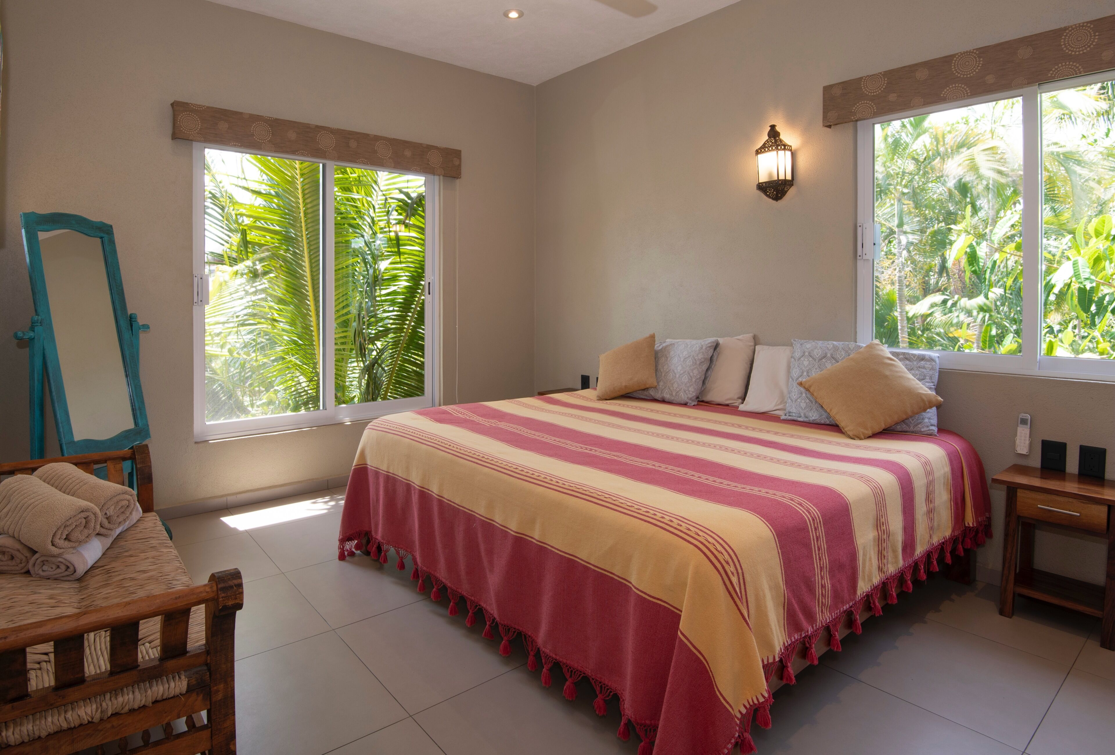 Une chambre avec un grand lit, deux fenêtres donnant sur la verdure, une chaise en bois et une table de chevet avec une lampe.
