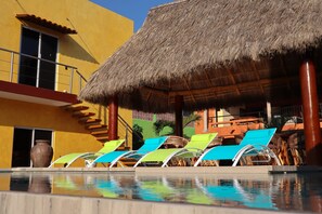 Un espace piscine avec des chaises longues et une structure dotée d’un toit de chaume.