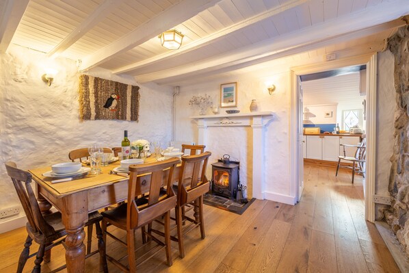 A dining area with a wooden table set for six, a stone fireplace, and a kitchen visible through an open doorway. - A spacious, detached cottage with character and charm, located in St Agnes. (St Agnes)