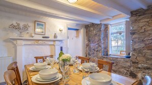 A dining table set with white plates, bowls, and cutlery, surrounded by wooden chairs, with a stone wall and a window in the background. - A spacious, detached cottage with character and charm, located in St Agnes. (St Agnes)