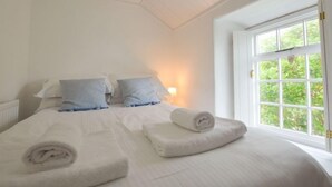 A bedroom with a large bed, two pillows, a bedside lamp, and a window with a view of greenery. - A spacious, detached cottage with character and charm, located in St Agnes. (St Agnes)