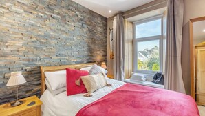 A bedroom with a stone wall, a large window with curtains, a bed with multiple pillows, and a bedside table with a lamp. - 2 bedroom accommodation in Windermere (Windermere)