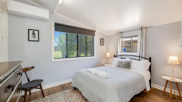 Un dormitorio con cama, lámparas de noche, ventana con vista a árboles, escritorio con silla y aire acondicionado instalado en la pared.