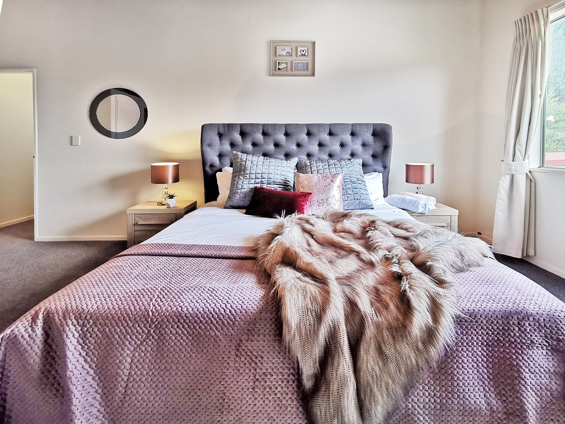A bedroom with a tufted headboard, a large bed with a patterned bedspread, multiple pillows, bedside tables with lamps, and a framed picture on the wall.