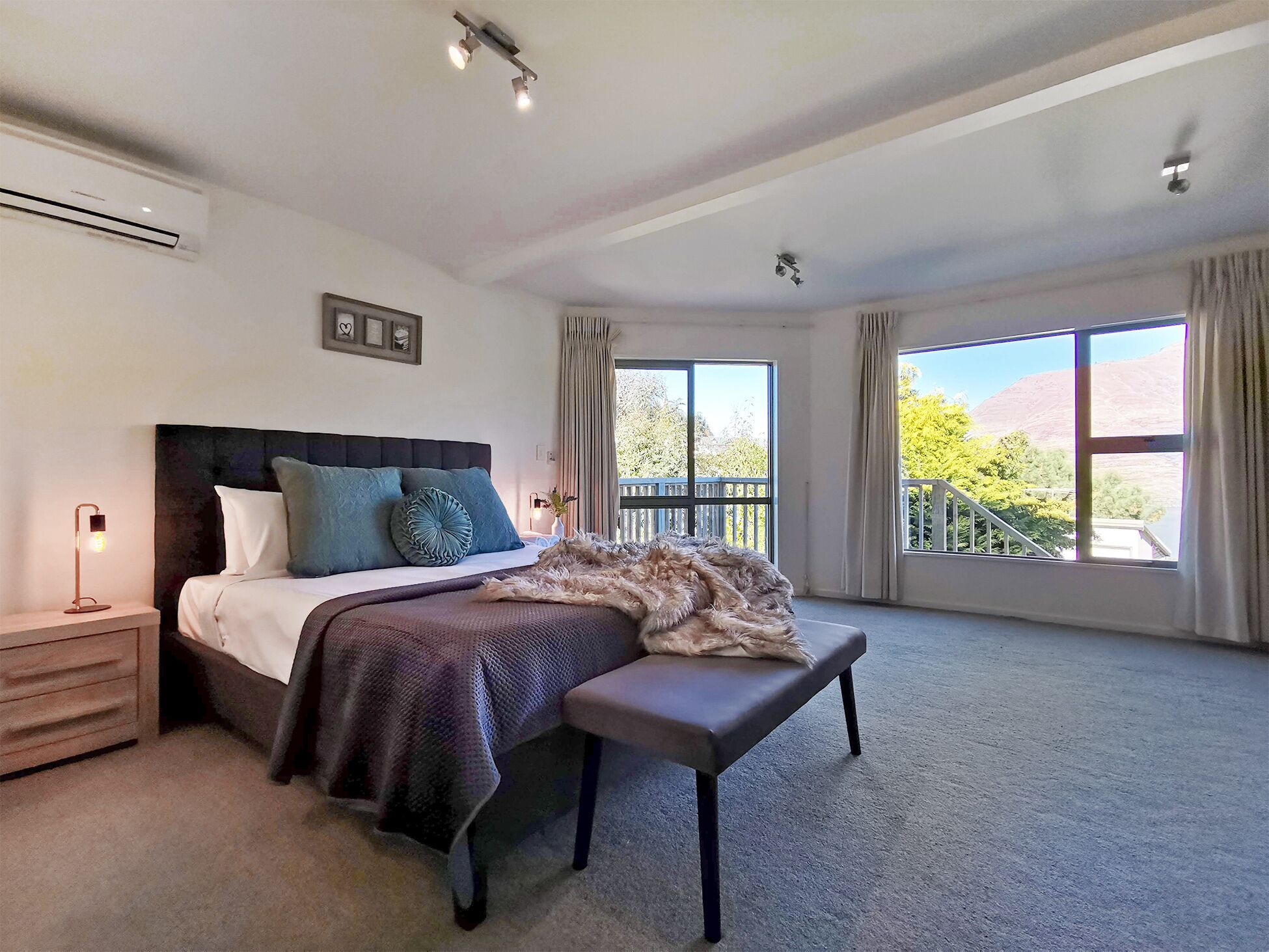 A spacious bedroom with a large bed, a bench, a nightstand, and a window with a view of trees and hills.