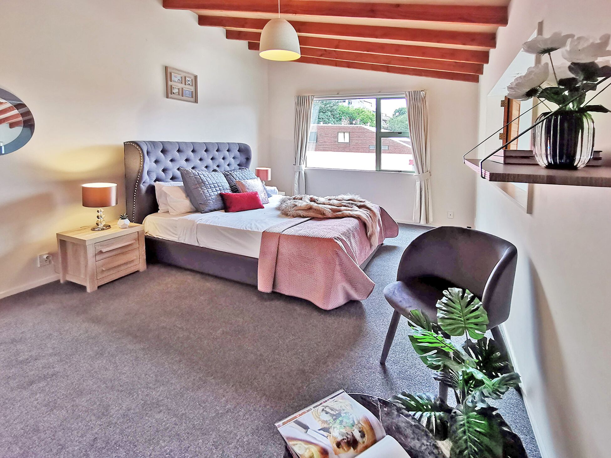 Une chambre à coucher avec un grand lit, des tables de chevet, une chaise, une fenêtre avec des rideaux et une plante en pot.