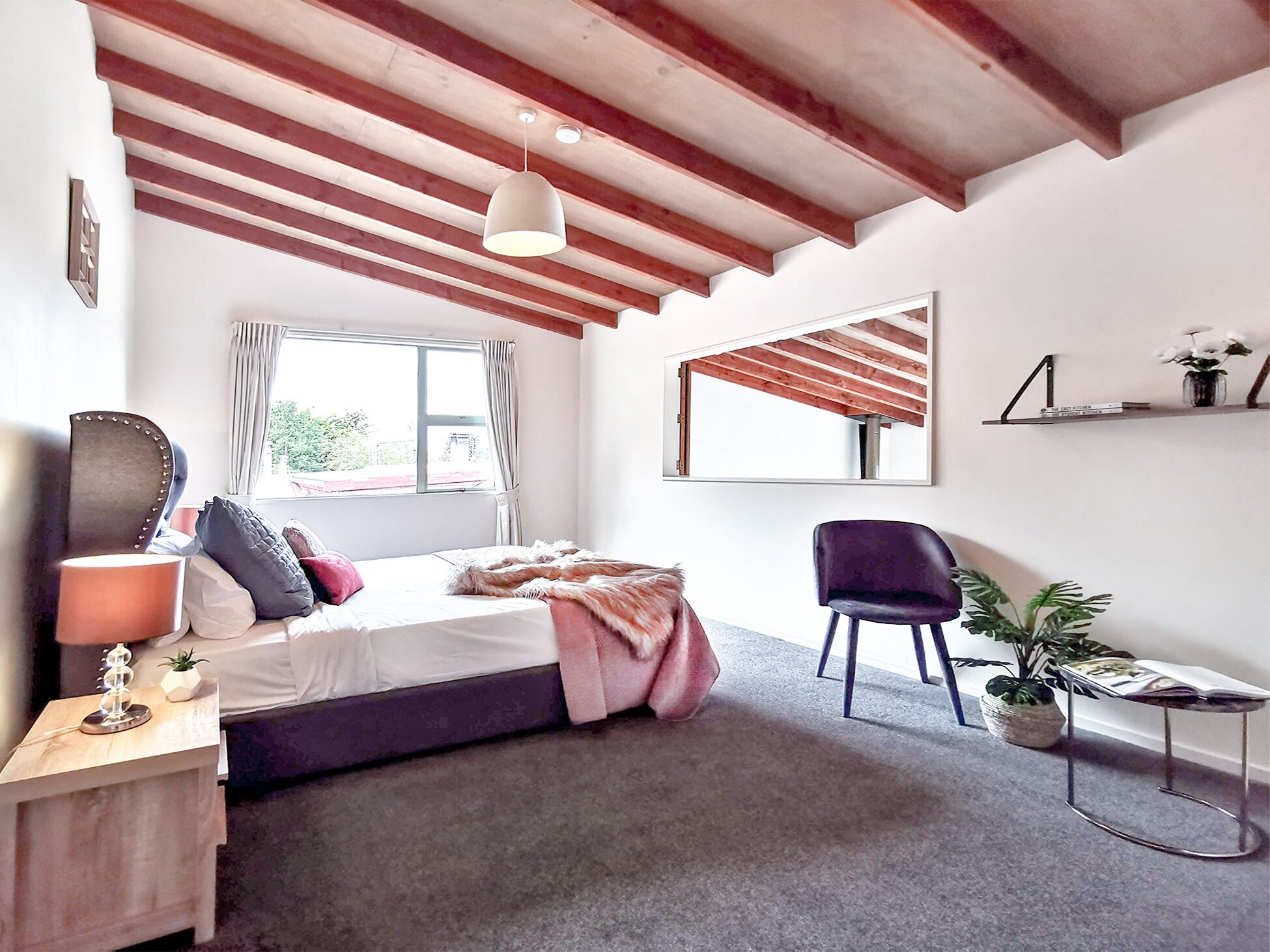 A bedroom with a bed, a chair, a bedside table, a mirror, a window, and a ceiling with exposed beams.
