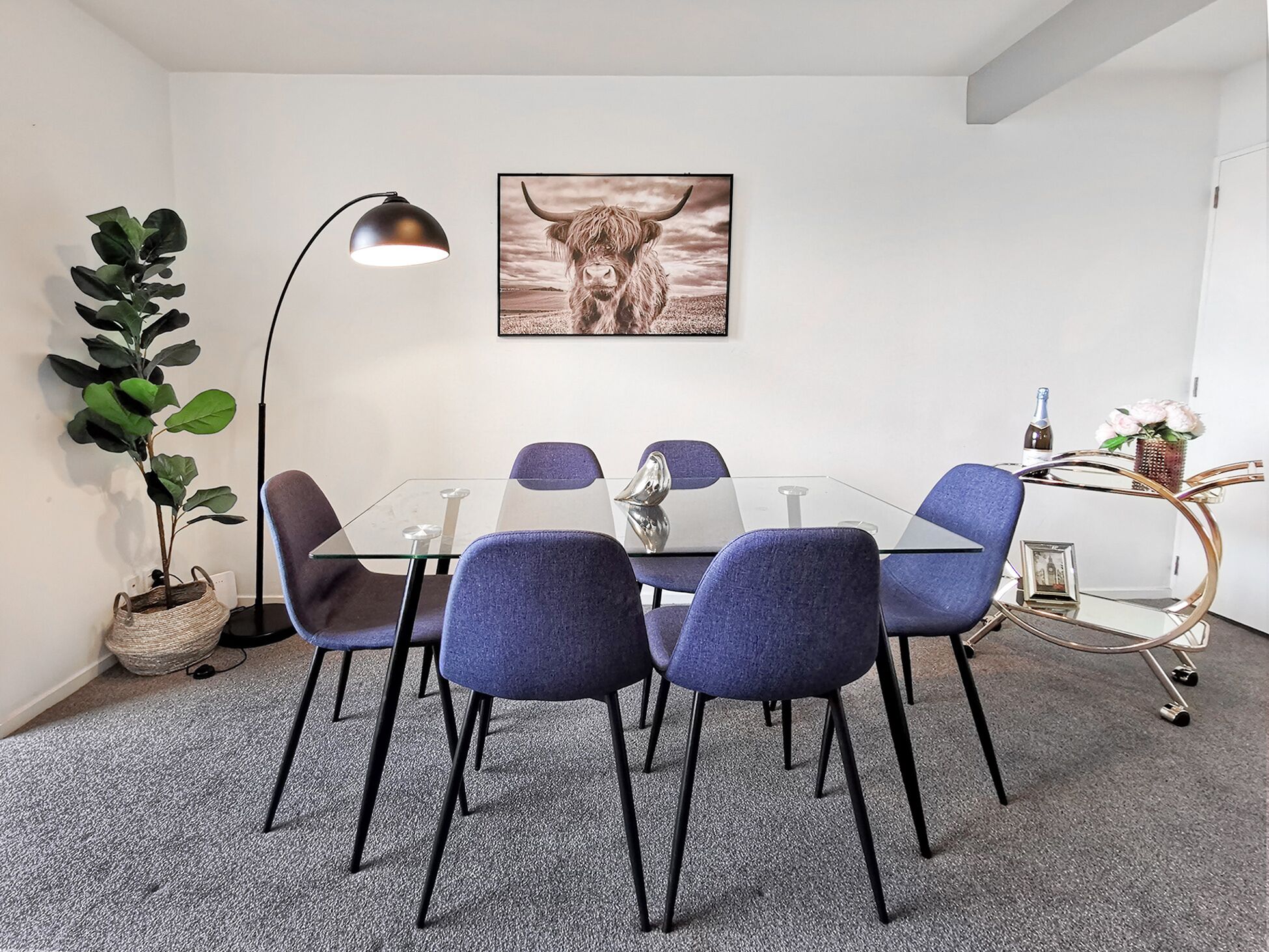Et moderne spiseområde med et glasbord, fire blå stole, en gulvlampe, en potteplante og et indrammet kunstværk på væggen.