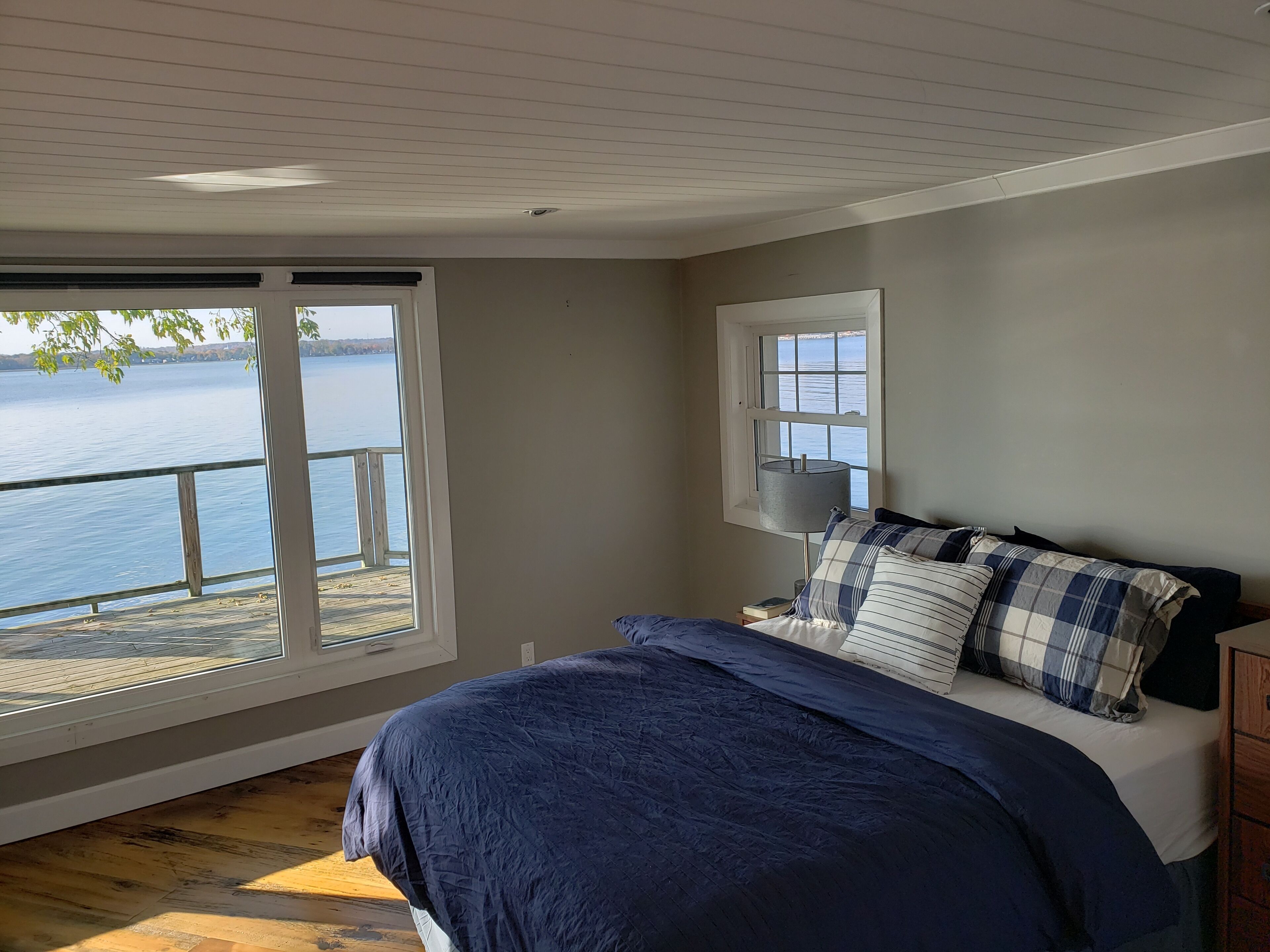 A bedroom with a large window overlooking a body of water, a bed with blue bedding, and a small bedside table.