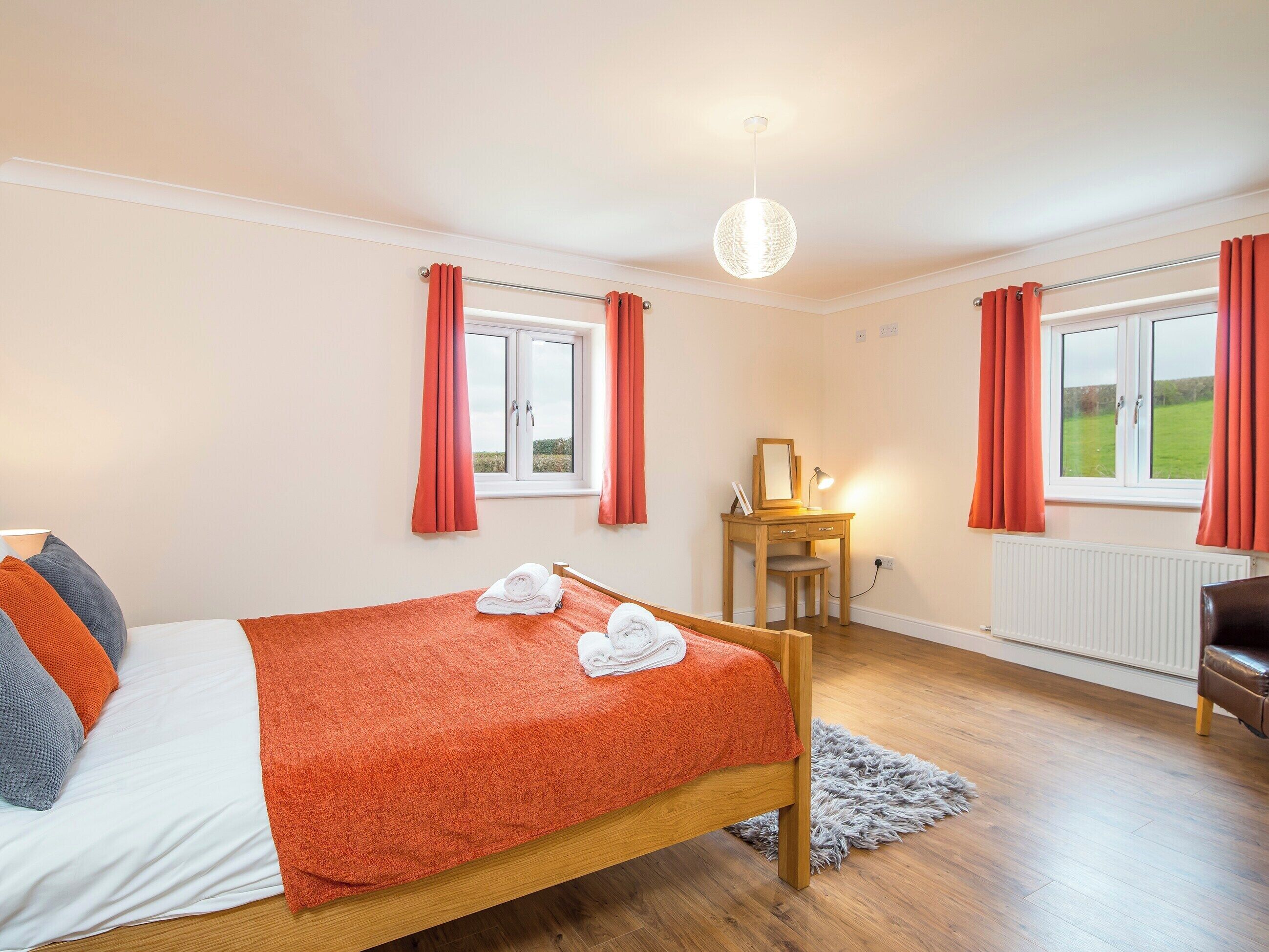 A bedroom with a wooden bed, a desk with a mirror, and two windows with red curtains.