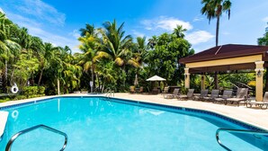 Une piscine d’un bleu éclatant, bordée de carreaux, entourée de chaises longues et d’un espace salon abrité, avec des palmiers en arrière-plan.