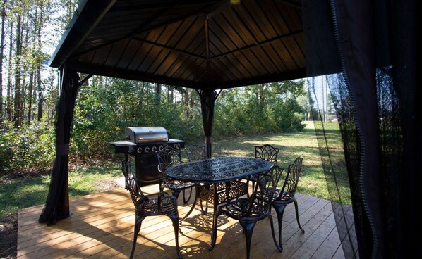 Enjoy al fresco dining on the patio, where a round table and four chairs await, accompanied by a barbecue grill and a shaded pergola amidst lush greenery.