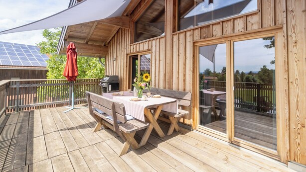En træterrasse med et bord og stole, en rød parasol og solpaneler i baggrunden.