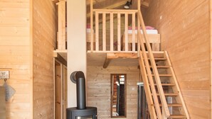 A wooden cabin interior with a central wood-burning stove, a loft bedroom with a bed, and a staircase.