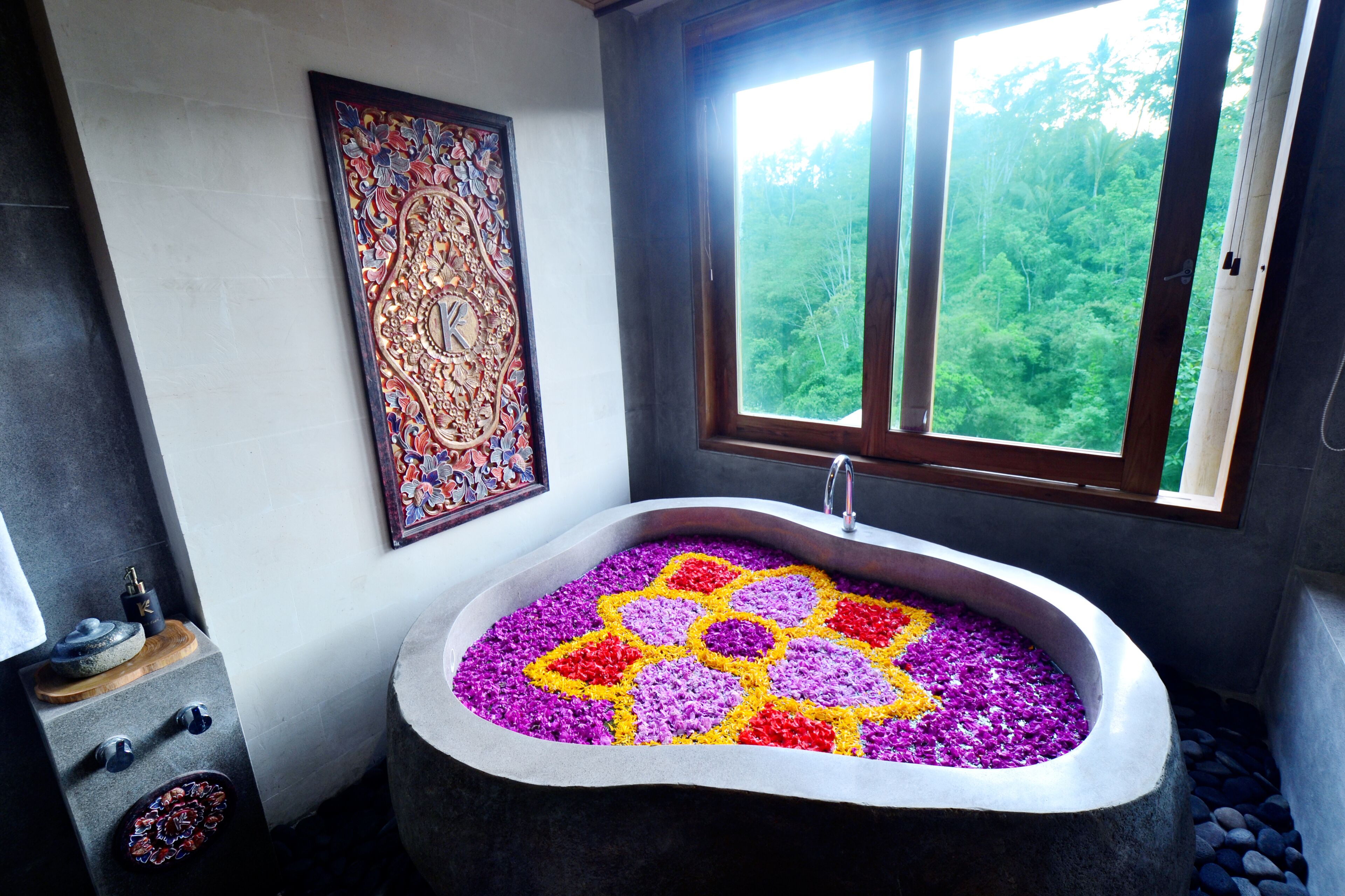 A bathroom with a large bathtub filled with colorful flower petals, a window with a view of greenery, and a decorative wall piece.