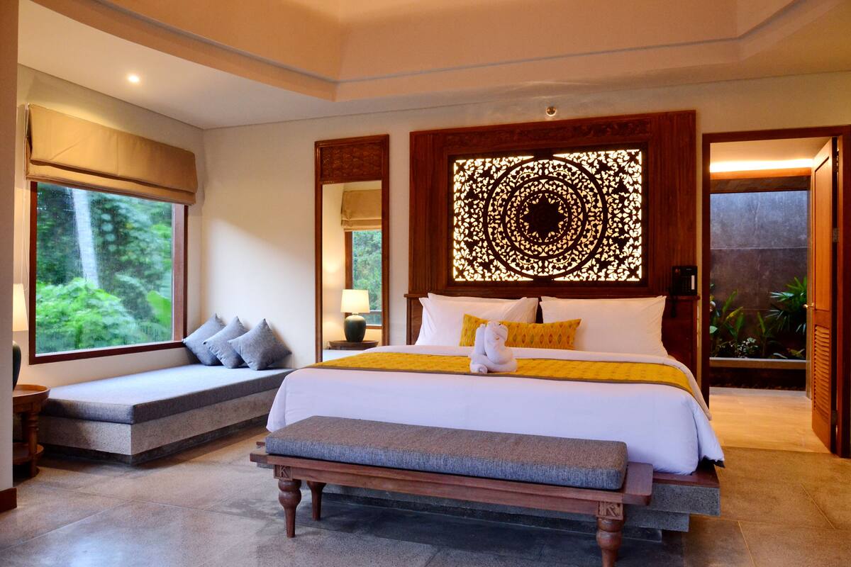 A spacious bedroom with a large bed, a bench, and a window with a view of greenery.