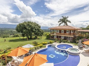 Area piscina con vista sul paesaggio, un edificio con balcone e arredi esterni ombreggiati.