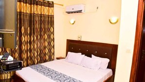 A hotel room with a bed, a bedside table with a remote control, an air conditioning unit, and patterned curtains. - Hôtel Kin Comfort (Kinshasa)