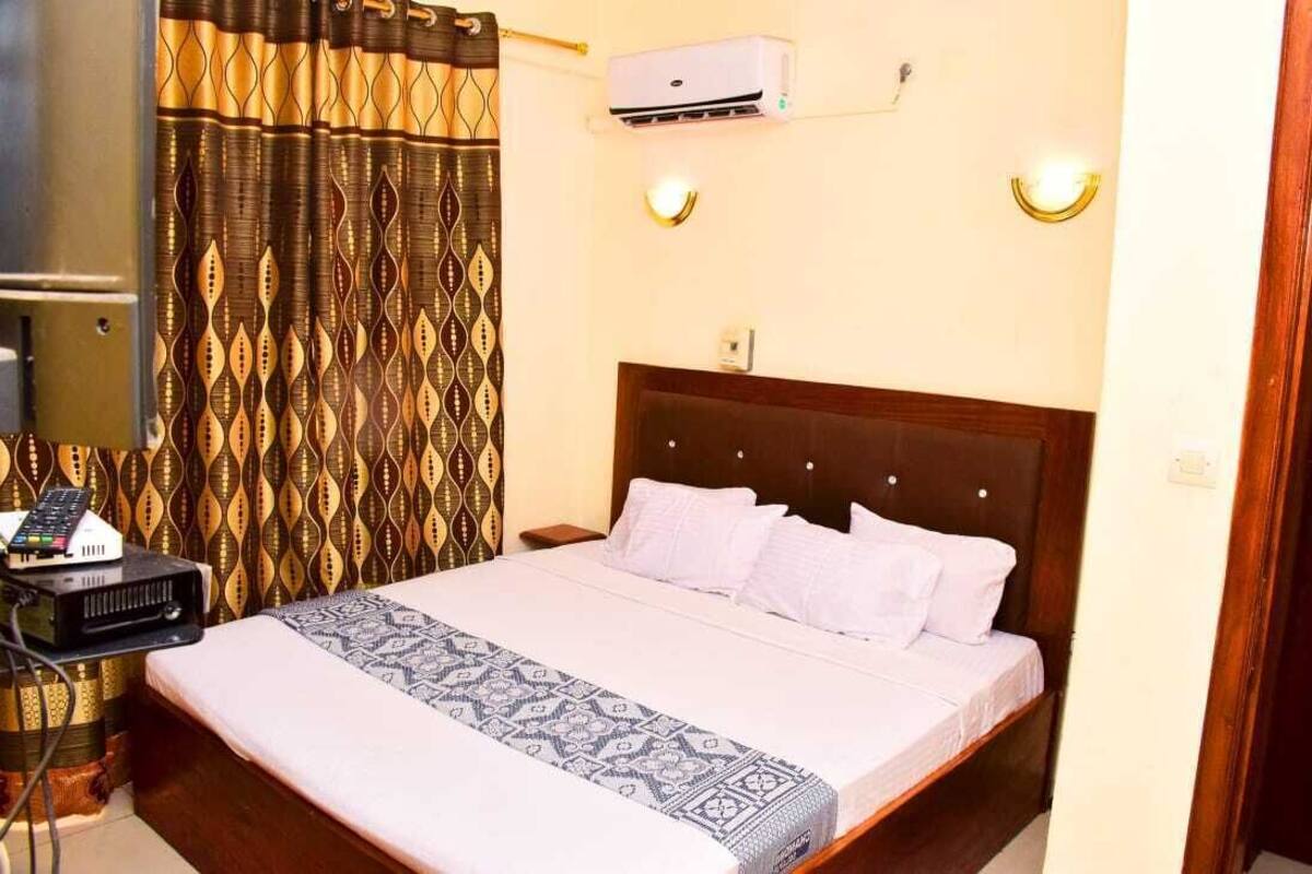 A hotel room with a bed, a bedside table with a remote control, an air conditioning unit, and patterned curtains.
