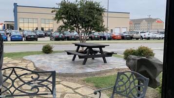 An outdoor patio area with a picnic table and metal chairs, a small tree, and a parking lot with various cars and a hotel in the background.