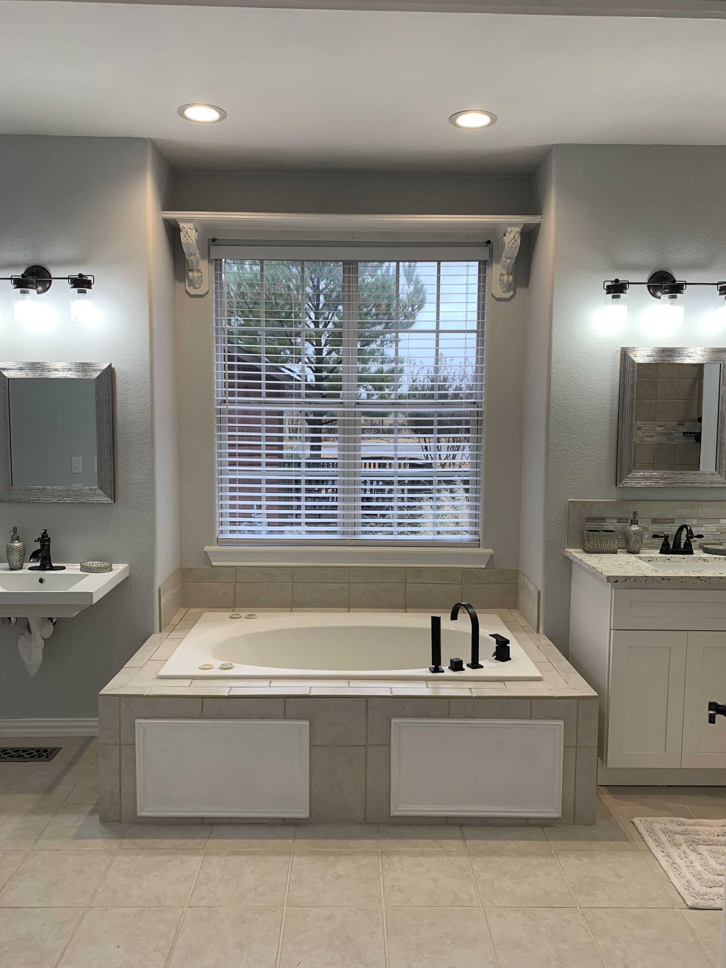 A modern bathroom with a large bathtub, two sinks, and a window with blinds.