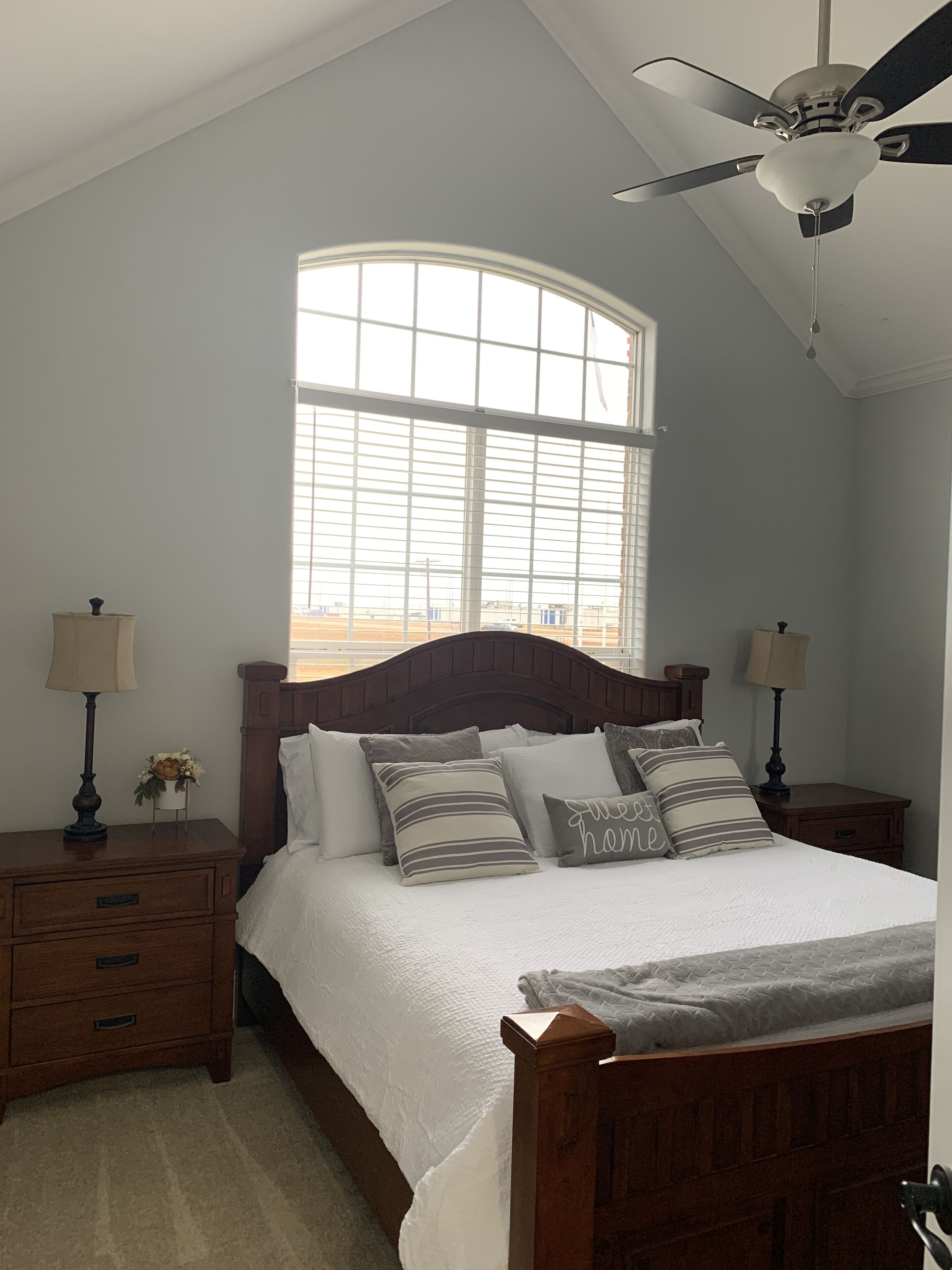 A bedroom with a large window, a bed with multiple pillows, two bedside tables with lamps, and a ceiling fan.