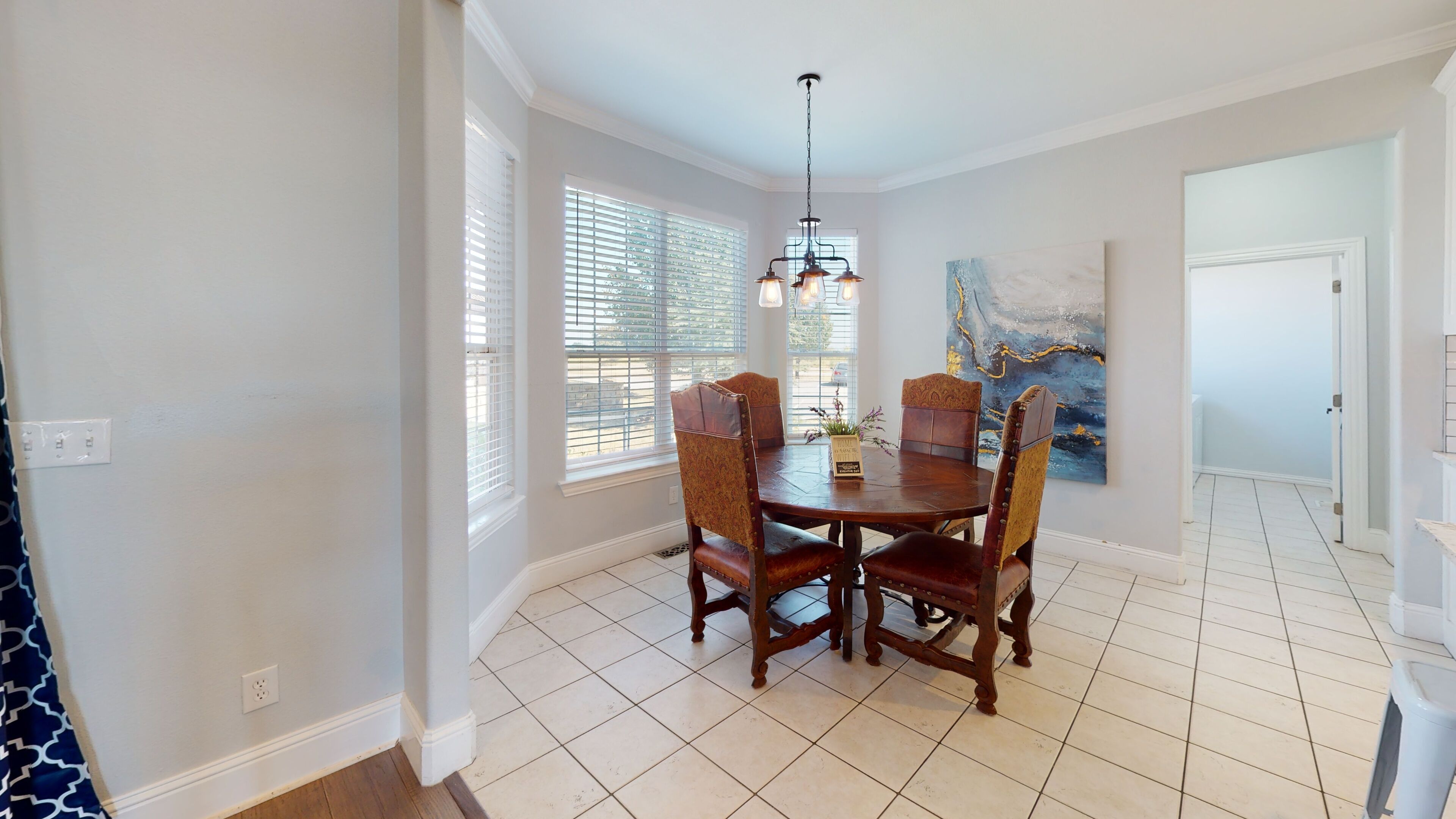 A dining room with a wooden table and chairs, large windows with blinds, a chandelier, and a painting on the wall.