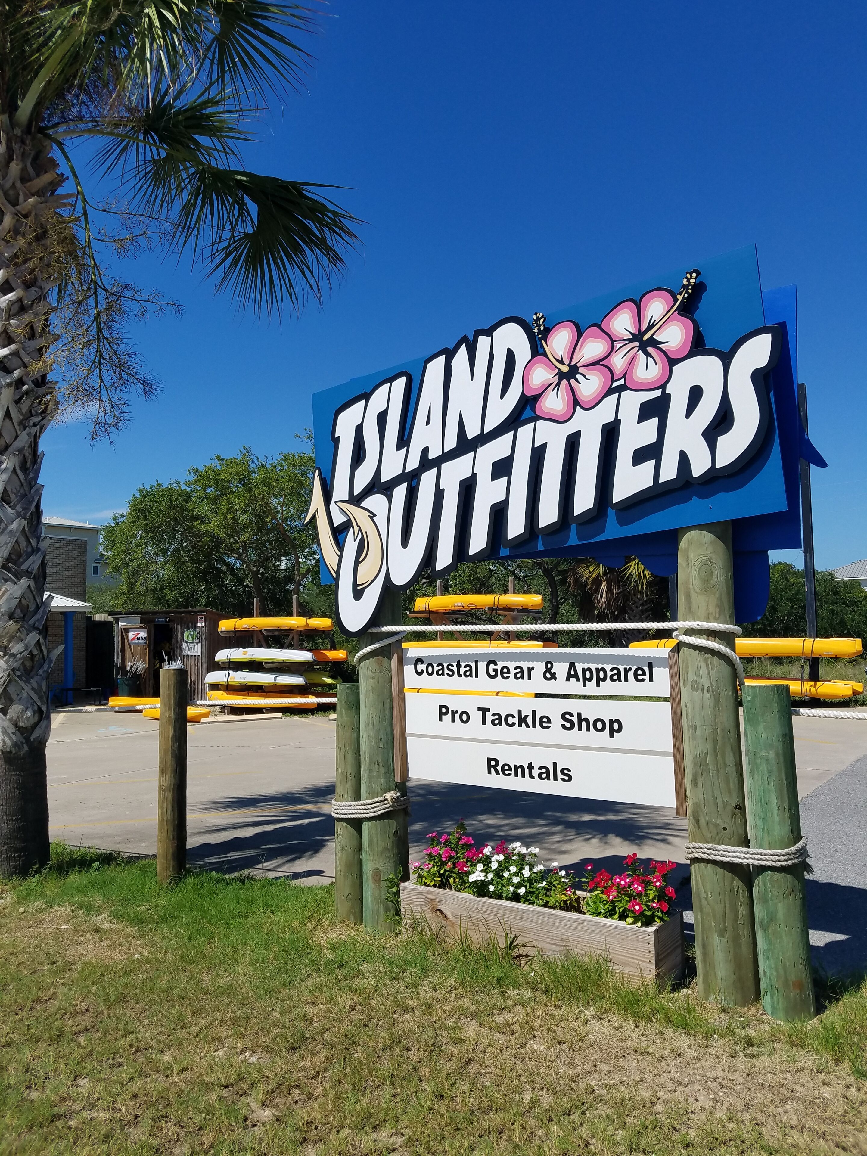 A sign for 'Island Outfitters' with a floral design, listing coastal gear, apparel, tackle shop, and rentals.