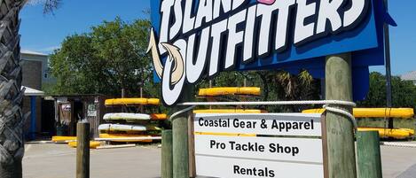 A sign for 'Island Outfitters' with a floral design, listing coastal gear, apparel, tackle shop, and rentals.